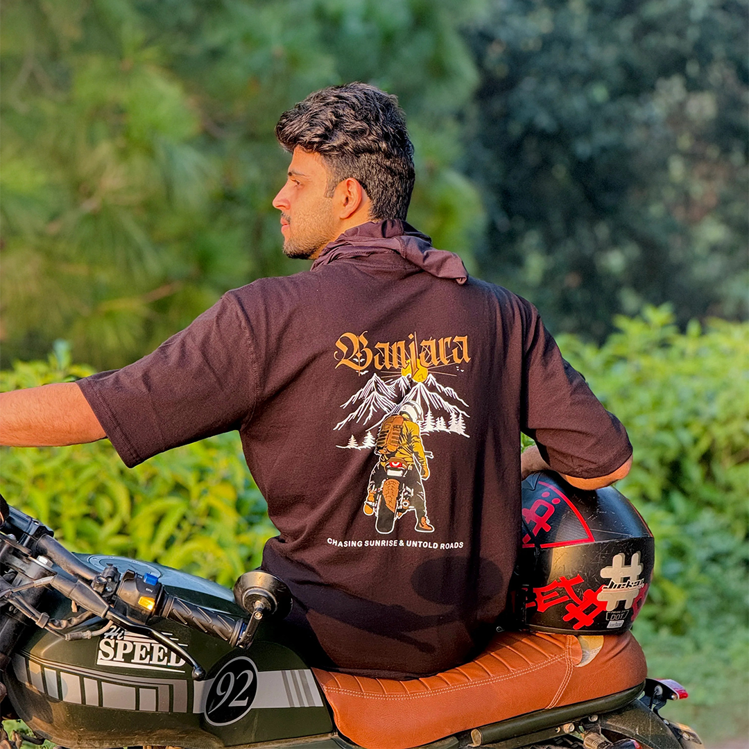 A man wearing a dark brown t-shirt with a large graphic print on the back, sitting on a motorcycle outdoors. The shirt features a graphic of a warrior in front of mountains and the text 'Vanijra'.